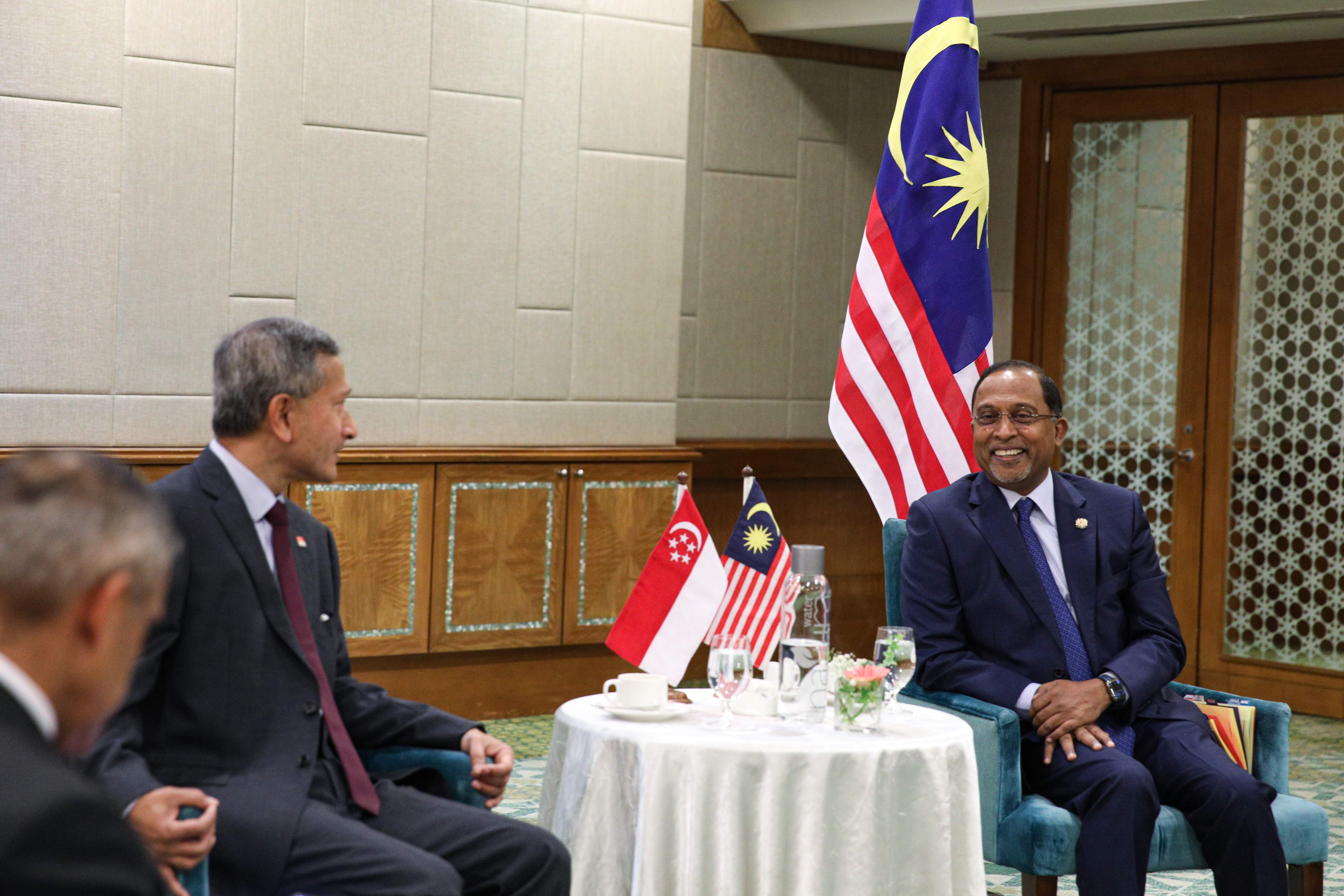 Two men in suits sit near flags of Singapore and Malaysia at a meeting.